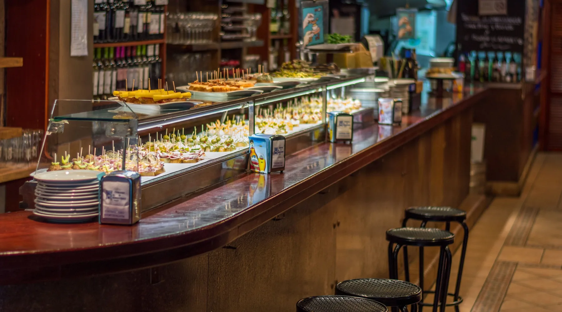 A long wooden bar counter with a glass case of small pinchos on skewers and plates, wine bottles on shelves behind, and black stools along the front.