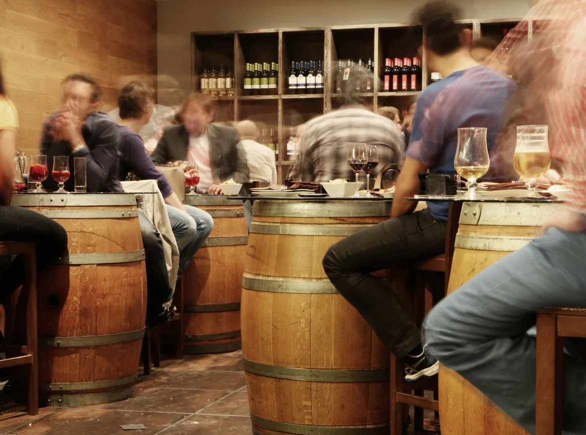A busy bar interior where people sit on stools around barrel tables with glasses of wine and beer, bottles lined on wooden shelves behind.