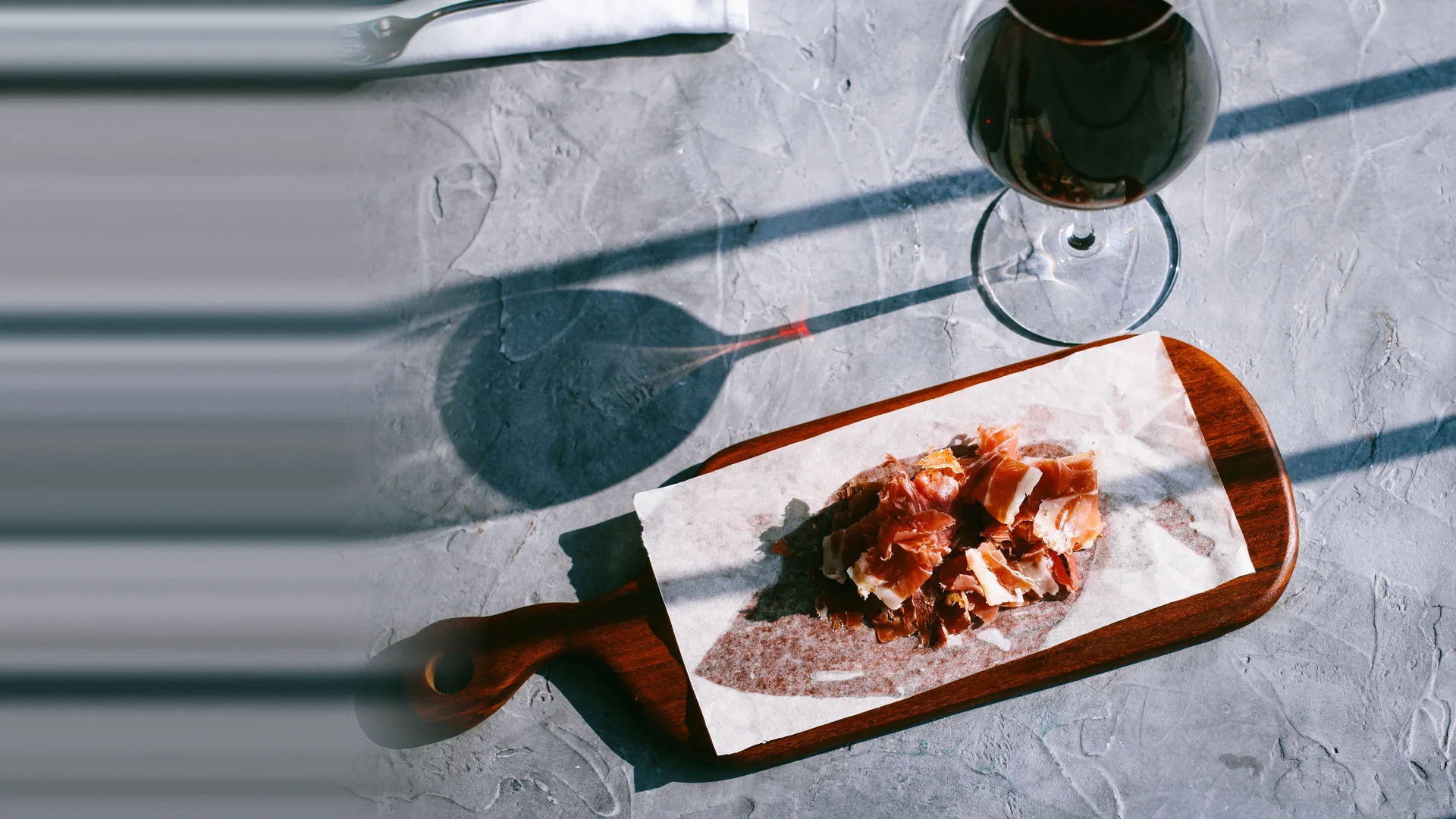 Slivers of cured ham on paper over a wooden serving board, with a glass of red wine beside it, on grey stone in strong sunlight.