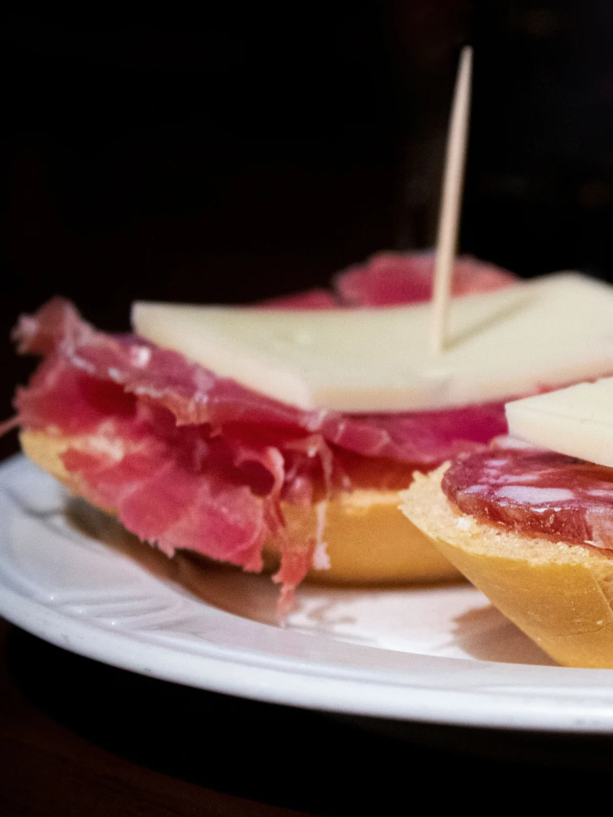 Two small open sandwiches on a white plate, one topped with cured ham and a slice of pale cheese, the other with salami and cheese, each held with a toothpick, against a dark background.