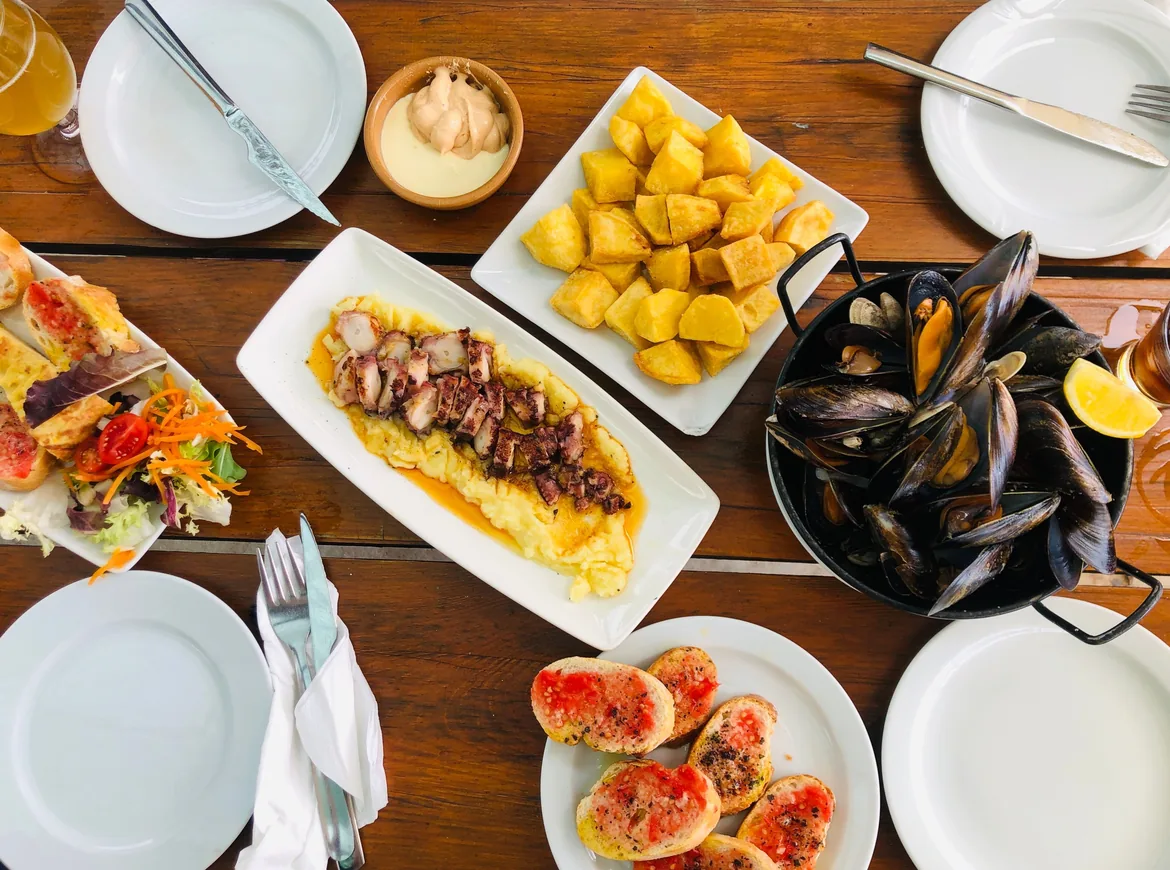 A wooden table seen from above set with fried potato cubes with a dip, sliced octopus on potato purée, a pan of mussels, tomato toasts and a glass of beer.